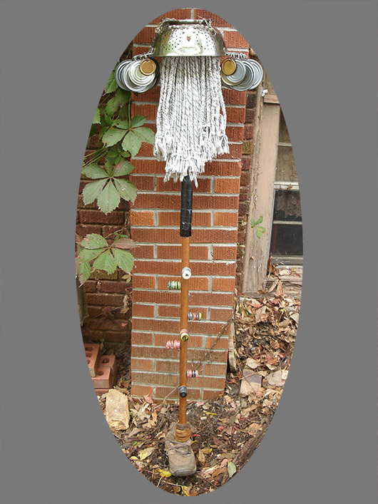 Full-length view of Mendoza with colander head, and bottlecap jingles nailed to mop stick, which stands in an old boot.