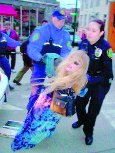 Lady Passion in blue tie-dye skirt having her arms twisted behind her back and being dragged down by two police officers
